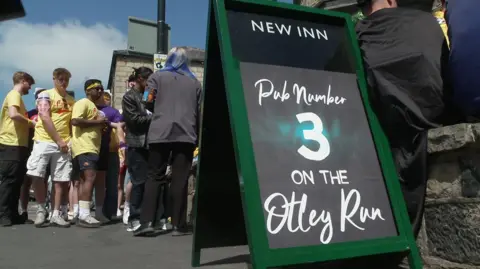 A sign on a public street reads: "New Inn; Pub number 3 on the Otley Run". Some people, including some dressed in yellow t-shirts mill about in the background.