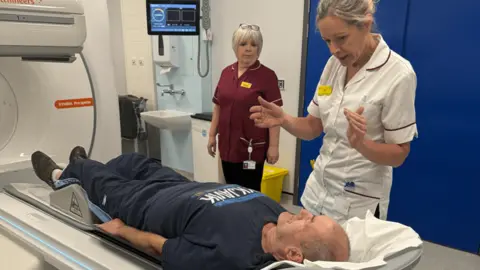 The Shrewsbury and Telford Hospital NHS Trust Two women in medical uniform stand by a man lying on a scanner unit.