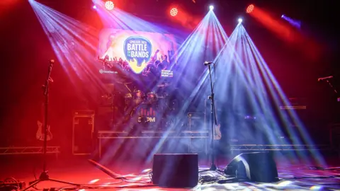 A stage set with drums, microphones, speakers and lit in red and blue. A sign at the back reads UNDER 18S BATTLE OF THE BANDS.