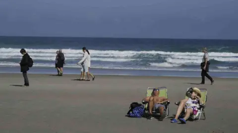 EPA A man and a woman lying on a chair on a beach with people walking and waves breaking behind them