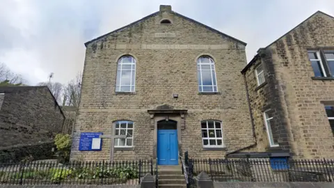 A square stone building with a blue door, four windows and Methodist Church inscribed on the front in stone. A blue sign on the left hand side, a cross standing in the flowerbed in front of the building. 