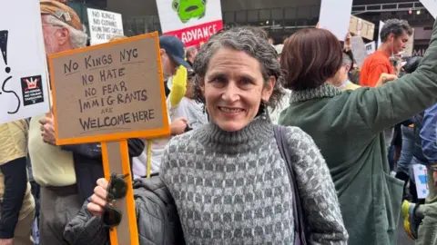 Grace Eliza Goodwin/BBC Beth smiling at camera in front of crowd of protesters, holding a sign that says