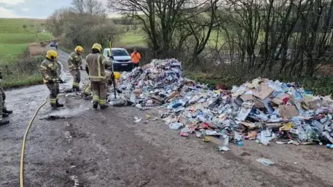 DORSET COUNCIL Three fire officers  stand next to two large piles of rubbish on the road. One of them is pointing a fire house at one of the piles which has been recently extinguished