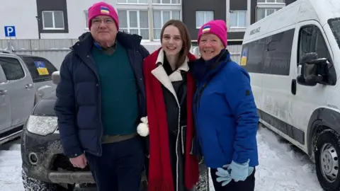 A man on the left wears a green jumper and a navy puffer jacket. He is also wearing a pink hat with a Ukraine flag on it. A woman in the middle wears a dark jacket and a red scarf. A woman on the right wears a blue jacket, blue gloves and a pink hat with a Ukraine flag on it. It is a snowy setting. Vehicles and an apartment are in the background.
