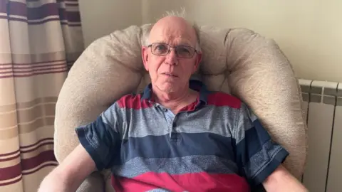BBC Charles is looking at the camera, wearing glassed and a striped polo shirt while sat in an armchair in front of a white wall with electric radiator next to the edge of a burgundy, champagne and cream striped curtain hanging down on his right side.