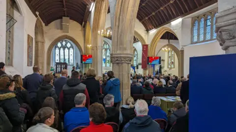 A busy church in Yate, packed with people both standing and sitting. They are looking towards the front of the building, and a picture of poppies is visible on the screens. 