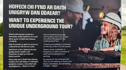 A sign telling visitors that Amgueddfa Cymru will be charging for underground tours at the Big Pit Musuem due to financial pressures. The sign has a background picture of a man and a young girl wearing white hard hats in an underground tour