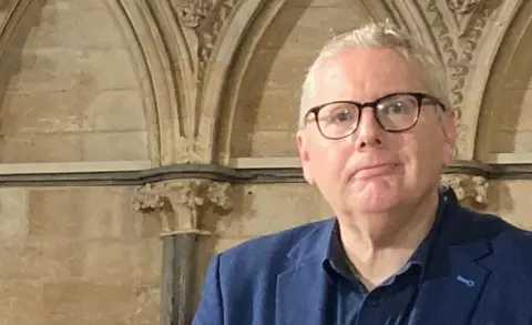 Tim Iredale with short light hair, dark-rimmed glasses and a dark blue suit and shirt stands in front of a medieval stone wall