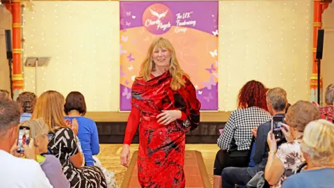 Ian Metcalf Model Colmer Hill on the catwalk during the 2023 show. She is smiling and the audience is clapping or taking pictures with smartphones. She is wearing a red and black dress.