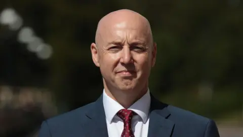 PA Media Jon Burrows is bald and is wearing a dark grey suit jacket with a white shirt and red tie