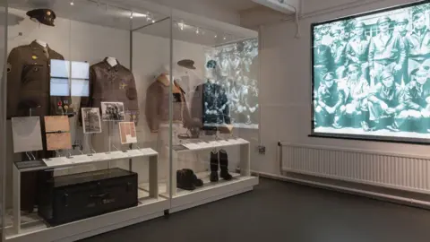 IWM Duxford The glass display case in the Air Crew Room at RAF Duxford. There are four uniform jackets on display, including a leather flying jacket. There are also photos, documents and footwear. On the right is a screen showing a black and white photo of at least 10 US aircrew. 