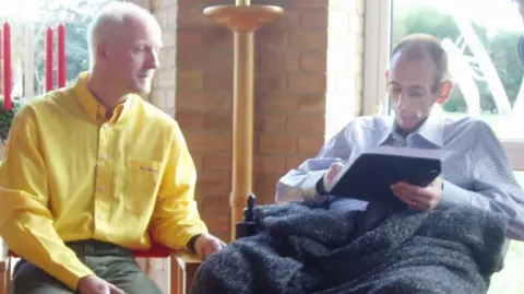 Chris Cramp A man in a yellow shirt, sat left, and a man wearing a light coloured shirt, sat right in a wheelchair. The man on the right is holding a black clipboard with a piece of paper on top of it.