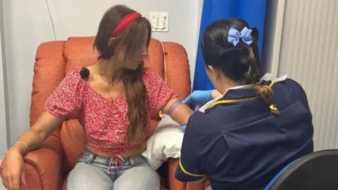 A woman sat on a hospital chair having her blood taken from her arm by a nurse