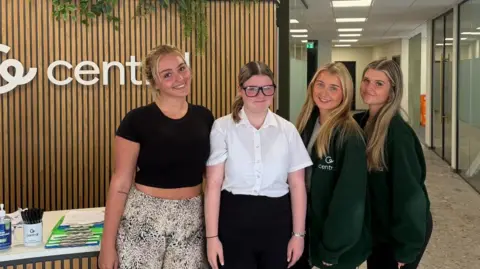 Central Employment North East Four people stand beside a reception desk in a modern office. Behind them is a wooden feature wall with a company logo and plants, while glass-walled meeting rooms line a corridor to the right.