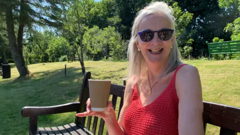 A woman in a red vest top with long blonde hair and sunglasses holds up her coffee cup while sitting on a park bench in among lawns and trees