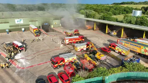 Mike Sudds An aerial of the waste recycling centre where fire service crews are parked up. There are firefighters on the ground and smoke is coming from one area of the building. Another part of the site looks blackened with smoke damage.