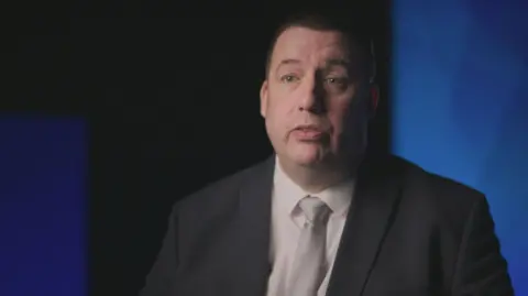 BBC Crimewatch DCI Ben Lavender wearing a white shirt, light grey tie and black suit. He is sitting in front of a blue background, giving an interview for BBC Crimewatch.