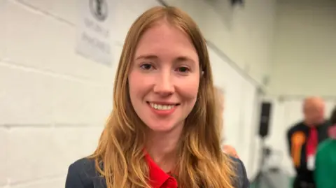 A woman with long, ginger hair smiling at the camera. She's wearing a red shirt and grey jacket, and standing in front of a white wall