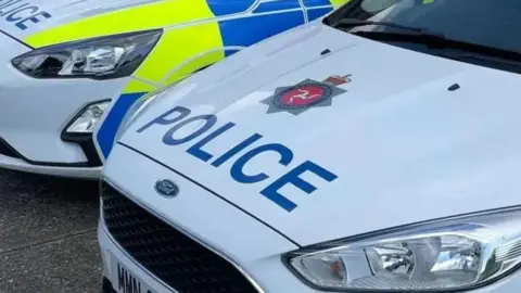 BBC The bonnets of two police cars parked next to each other. The nearest has the word police in blue writing on a white background, and features the Isle of Man Constabulary's emblem. Blue and yellow paint can be seen on the side of the other car.