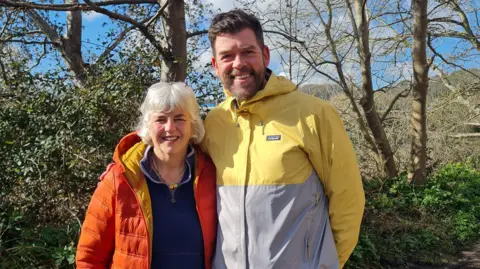 Bristol Walk Fest A man and a woman stand alongside each other looking at the camera. She has white hair and is wearing an orange padded jacket. He has a beard and is wearing a waterproof top which is a mixture of yellow and grey in colour. They are part of the Bristol Walk Fest