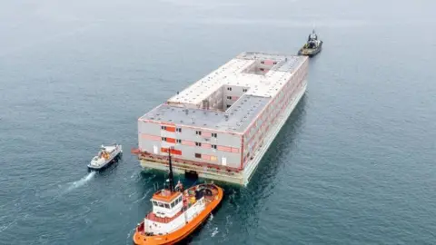 PA Media A small orange boat next to a large barge in the sea.