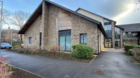 Lanchester Medical Centre is a beige brick building with a large triangle roof. It has large windows with Lanchester Medical Centre written across it with grey metal letters.
