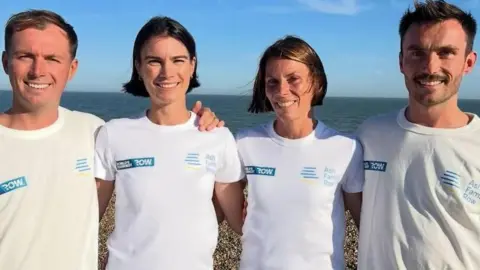 Two men and two women stand on a pebbled beach wearing matching white “World’s Toughest Row – Ash Family Row” T‑shirts, with the sea and blue sky behind them.