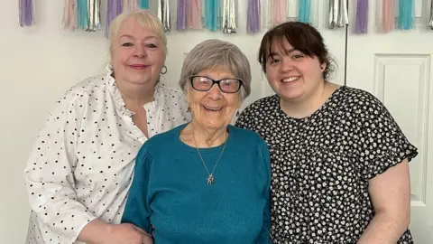 Anne is standing in the middle of her daughter Donna and her granddaughter Sarah. Anne has short grey hair and is wearing dark rimmed glasses, a blue jumper and a gold necklace and earrings. Donna has short blonde hair and is  wearing a white blouse with black hearts on and gold hoop earrings. Sarah has dark hair and is wearing a floral dress.