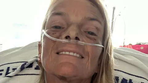 Mary-Jane in a hospital bed. She has blonde hair and a tube up her nose