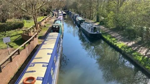 A view of a canal from a bridge. There's a series of brightly coloured narrow boats on either side and a reflection of the trees in the water.