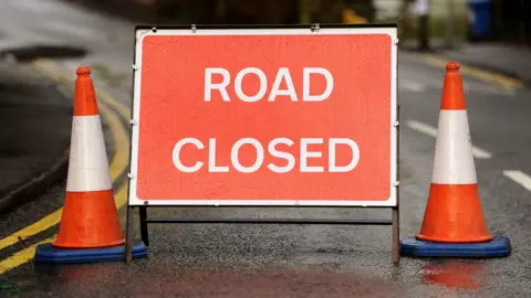 A red road closed sign with traffic cones either side