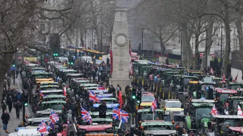 Gareth Fuller/PA Tractors lined up in four or five rows nose to tail and stretching as far back as the eye can see. They are parked on Whitehall in London, with the column of the Cenotaph war memorial in amongst them and reaching into the air. There are people milling around the tractors and there are union flags attached to several of them. It appears to be a wintry day and the trees on either side of the road are showing their bare branches.