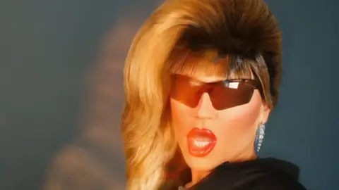 CAA A head shot of a drag queen - Jodie Harsh - with big blonde hair falling to one side, black sunglasses, long diamante earrings and her lips open, against a charcoal grey backdrop. 