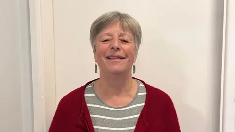 LDRS Sarah Hill is standing infront of a white door, smiling at the camera. She is wearing earings, a stripey top and red cardigan. 