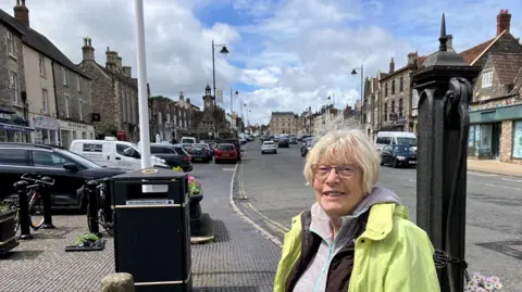 A woman with white hair and glasses is standing at the side of a road. She wears a lemon yellow jacket. To one side of her is a metal water pump, to the other a black public bin and a flag pole. Cars are parked in the distance as the road tapers towards the clocktower