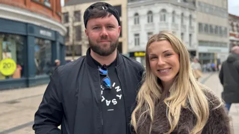 Alex and Kate smiling at the camera. Alex is wearing a black jacket, backwards baseball cap and a black tshirt with white writing. He has brown hair. Kate is wearing a brown fur jacket and has long blonde hair.