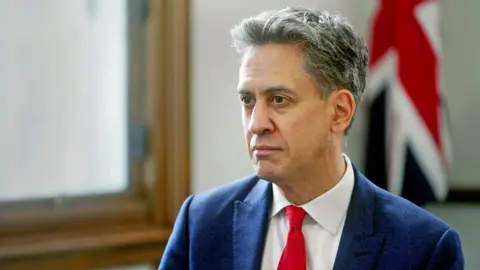 Ed Milliband depicted during his interview. He has short, black, brown hair. He is wearing a Navy suit, white shirt and red tie. He is behind a wooden window frame and a union jack flag. 