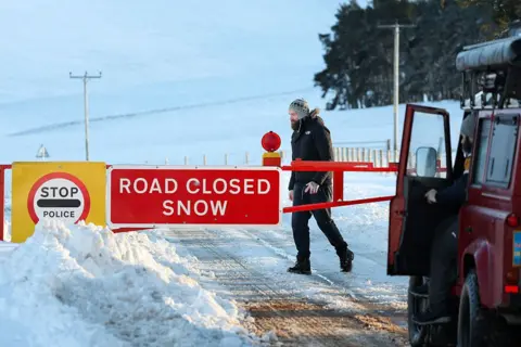 Getty Images Members of the public are seen at the snow gates at the A939 Cockbridge in Tomintoul, UK. The gate says 'road closed snow' on a red sign.