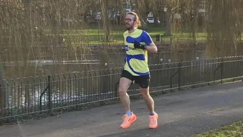 Chris White Chris White is running along a paved path in a park during daylight. He is wearing a bright yellow and blue athletic vest, dark shorts, and neon orange running shoes. His vest says 'MS Trust' in blue writing. The path is beside a black metal fence that borders a small lake. The water is lined with tall willow trees with long, drooping branches. 