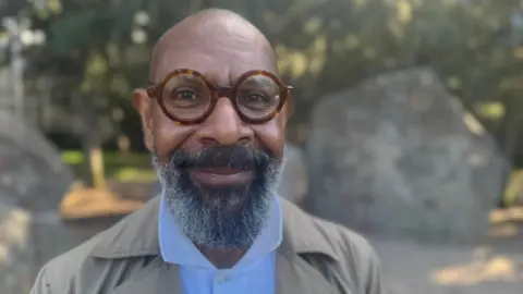 BBC/Simon Atkinson A man with brown rimmed glasses and a beard smiling at the camera while dressed in a light blue shirt and grey jacket. 