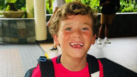 Alfie Kiely A young boy with curly blonde hair, wearing a red t shirt and black backpack and smiling at the camera. 