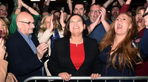 Getty Images Mary Lou McDonald celebrates with her fans after being elected to the RDS Count Center in 2020. He has a wide smile and is surrounded by cheering and clapping people.