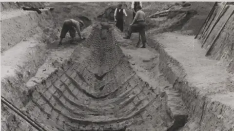 Mercie Lack/National Trust/PA The original burial sheep unearthed at Sutton Hoo. It is a black and white picture and shows the hull of the ship being excavated.