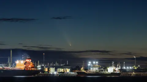 Playa Martin Cohen Warsach a las 19:30 GMT del 12 de octubre. En primer plano se ven varios barcos grandes y, a lo lejos, el cometa surca el cielo azul crepuscular.