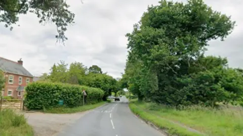 A screenshot from Google Streetview of a road in the countryside surrounded by trees and long grass. A brick house is on the left side of the road. 