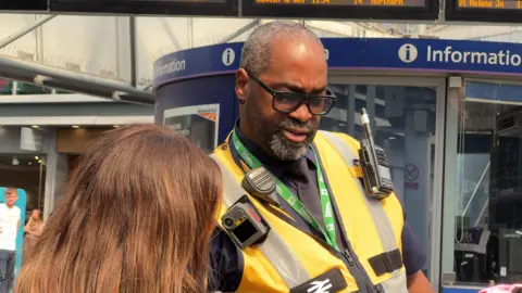 Network Rail A man with short grey hair and wearing black framed glasses, an orange high visibility vest with a radio, camera attached and a green lanyard is helping a passanger.