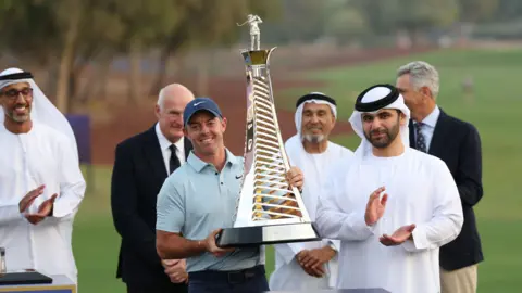 Rory McIlroy pictured holding a large trophy. On top of the trophy is a small statuette of Harry Vardon. Tournament officials are pictured in the background, along with a golf course.  