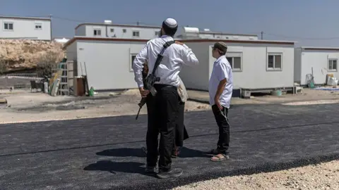Three Israeli settlers stand outside houses after the reopening of a settlement at Ganim in the Israeli-occupied West Bank on 13 August. One is a man armed with a rifle. The image has a blue border with the BBC Verify logo in the the top left corner. 