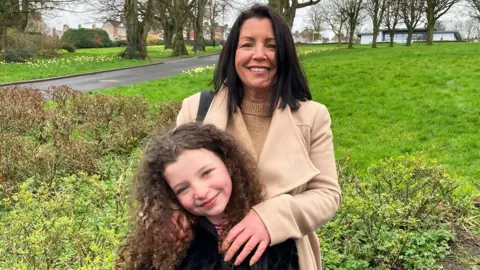Louise James stands behind her daughter Rioghnach. Louise has long dark hair and is wearing a tan woollen coat over a tan woollen sweater. She has her arms on Rioghnach who is smiling and has curly brown hair. They are standing in a park, trees and a rolling stretch of grass can be seen behind them.