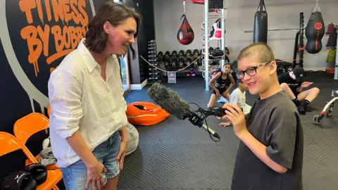 Mum Hayley is wearing a white short and demin shorts and is with her son Rex who is wearing a grey t-shirt. He has glasses and is holding a microphone.